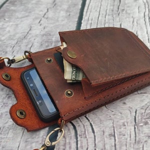 May include: A brown leather phone case with a strap. The case has a flap that can be closed with a snap. There is a small pocket on the front of the case that can be used to hold cash or cards.
