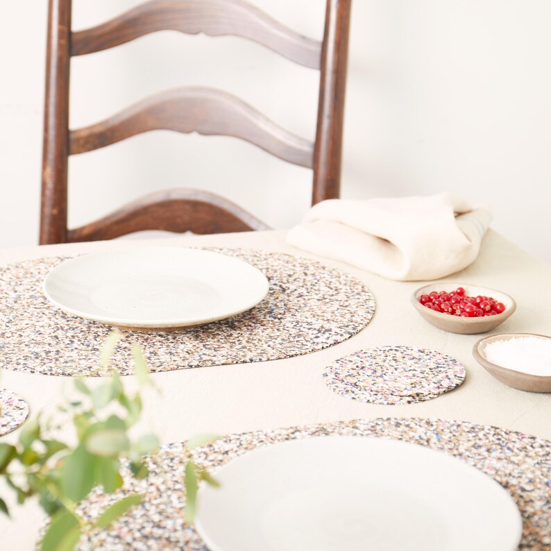 Peut inclure: Un ensemble de sets de table ronds et un dessous de verre assorti pr&eacute;sentant un motif mouchet&eacute; multicolore sur fond clair. Les sets de table sont dispos&eacute;s sur une table avec des assiettes en c&eacute;ramique blanche et deux petits bols.