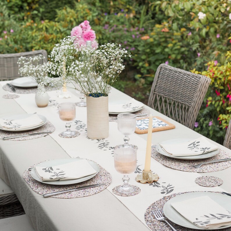 Peut inclure: Une table &agrave; manger dress&eacute;e avec un chemin de table beige &agrave; motif de feuilles sombres, des serviettes assorties, des sets de table ronds mouchet&eacute;s et des sous-verres. La table comprend des verres c&ocirc;tel&eacute;s, un vase cylindrique et une bougie.