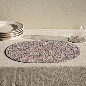 Peut inclure: Un set de table ovale et un dessous de verre rond assorti pr&eacute;sentant un motif mouchet&eacute; multicolore compos&eacute; de minuscules &eacute;clats noirs, blancs, bleus, roses et orange. L'ensemble est pr&eacute;sent&eacute; avec une pile d'assiettes cr&egrave;me et un verre.