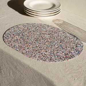 Peut inclure: Un set de table ovale pr&eacute;sentant un motif dense et multicolore compos&eacute; de minuscules taches dans des tons de noir, blanc, bleu, rose et orange. La surface poss&egrave;de une texture plate et uniforme avec un aspect confettis, offrant un contraste vibrant.