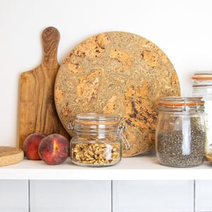 May include: A white shelf with a cork trivet, a wooden chopping board, a cork round serving board, two glass jars with lids filled with walnuts and chia seeds, a glass jar with a lid filled with white grains, two peach halves, and a stack of round cork trivets.