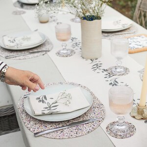 Peut inclure: Une table dress&eacute;e avec des sets de table ronds mouchet&eacute;s, un chemin de table blanc &agrave; motif de branches d'olivier noires, des assiettes gris clair, des serviettes pli&eacute;es &agrave; motifs floraux, des verres c&ocirc;tel&eacute;s et un grand vase cylindrique.