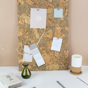 May include: A corkboard with a light brown and beige marbled pattern. The corkboard has several notes and cards pinned to it, including a light blue note with the text "Good vibes only", a white note with handwritten text, and a light pink card with a moon graphic. A green glass vase with dried flowers sits on a white surface in front of the corkboard. A white mug with a cork coaster sits on the right side of the image. A notebook with a colourful cover and white pages is open on the left side of the image.
