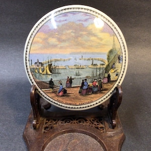 May include: A vintage ceramic lid with a colorful illustration of a harbor scene. The lid features a view of a harbor with boats, buildings, and people. The text "Royal Harbour Ramsgate" is printed on the bottom of the lid.