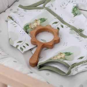 May include: A white baby onesie with a green trim and a woodland animal print. The onesie is lying on a white and gray polka dot crib mattress with a wooden teether shaped like a flower.