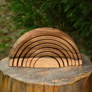 Pode incluir: Um brinquedo de empilhar de madeira em forma de arco-íris feito de madeira natural de cor castanho quente. O brinquedo é feito de sete peças que se encaixam e podem ser empilhadas para criar um arco-íris.