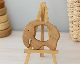 May include: A wooden elephant teether on a small wooden easel. The teether is light brown and has a hole in the middle. The easel is also light brown and has three legs.