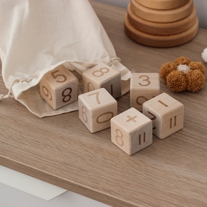 May include: Wooden blocks with numbers and a plus sign, in a white drawstring bag, on a wooden surface. There are also three pom-pom flowers in brown, white, and beige.