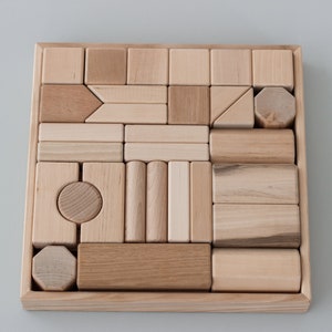 May include: A wooden box filled with various shapes and sizes of wooden blocks. The blocks are all natural wood color and are arranged in a grid pattern.