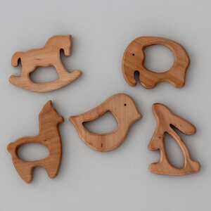 May include: Five wooden teethers shaped like animals: a rocking horse, a bird, a llama, an elephant, and a rabbit. The teethers are all made of natural wood and have a smooth, polished finish.