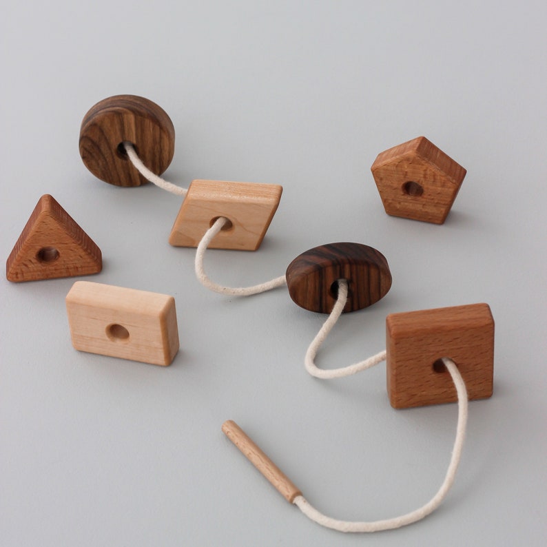 May include: A wooden toy with different shapes and a white string threaded through the holes. The shapes include a circle, a square, a triangle, a rectangle, and a pentagon. The toy is made of natural wood and has a rustic look.