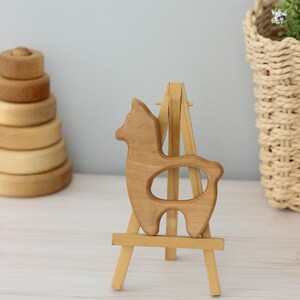 May include: A wooden llama toy on a small wooden easel. The llama is light brown and has a hole in the middle of its body.