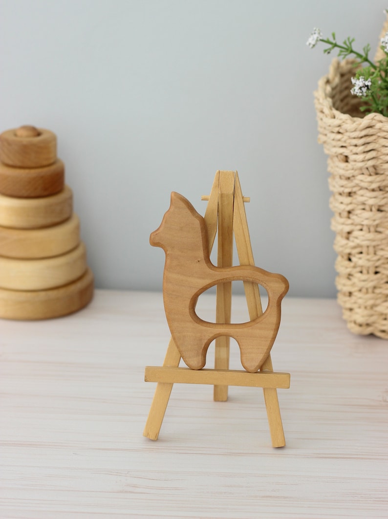 May include: A wooden llama toy on a small wooden easel. The llama is light brown and has a hole in the middle of its body.