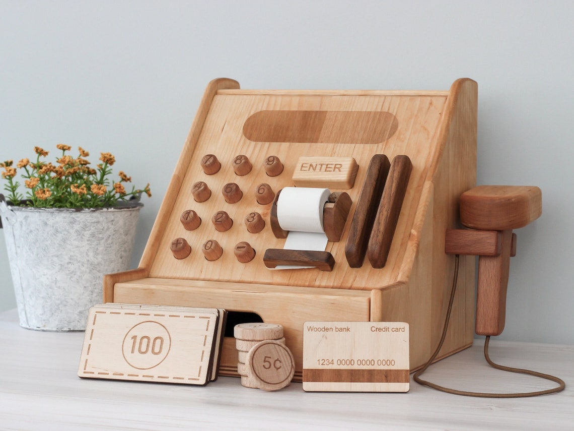 May include: A wooden toy cash register with a brown cord attached to a scanner. The register has a white paper roll, a wooden drawer, and a display with numbers 1-9 and an enter button. The register also has a credit card slot and a wooden bank with the text 'Wooden bank' and '1234 0000 0000 0000' printed on it. The register is on a light brown wooden surface.