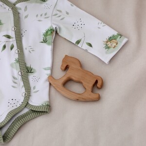 May include: A white baby onesie with a green leafy pattern and a small rabbit illustration. The onesie has a green trim and poppers down the front. A wooden rocking horse toy is lying on a beige surface next to the onesie.