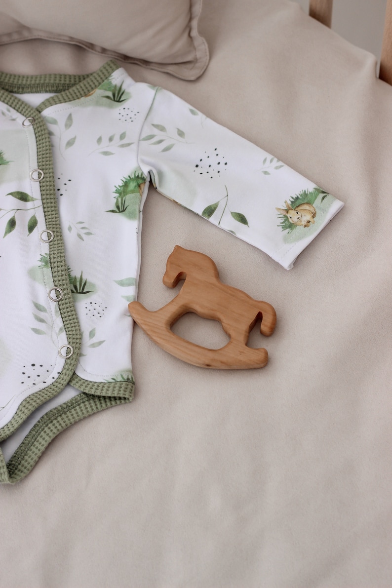 May include: A white baby onesie with a green leafy pattern and a small rabbit illustration. The onesie has a green trim and poppers down the front. A wooden rocking horse toy is lying on a beige surface next to the onesie.