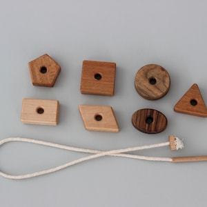 May include: A set of wooden shapes with holes in the center, strung on a white cord. The shapes include a circle, a triangle, a square, a rectangle, a pentagon, and an oval. The shapes are made of different types of wood, including light and dark wood.