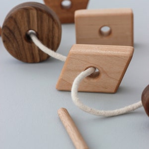 May include: A wooden toy with different shaped pieces strung together with a white cord. The pieces include a circle, a square, a diamond, and a pentagon. The toy is made of natural wood and has a rustic look.