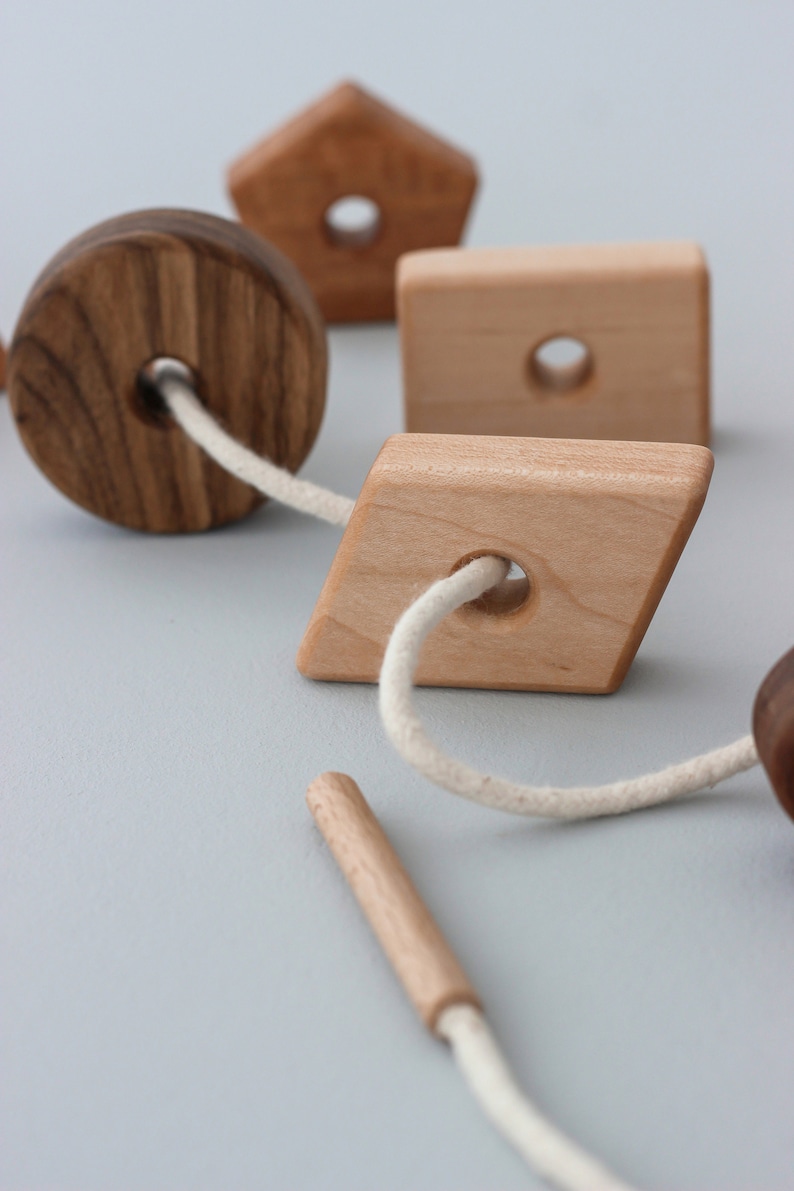 May include: A wooden toy with different shaped pieces strung together with a white cord. The pieces include a circle, a square, a diamond, and a pentagon. The toy is made of natural wood and has a rustic look.