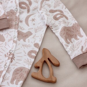 May include: A white baby onesie with a brown and white woodland animal print. The onesie has a snap closure and long sleeves. A wooden bunny teether is lying on the onesie.