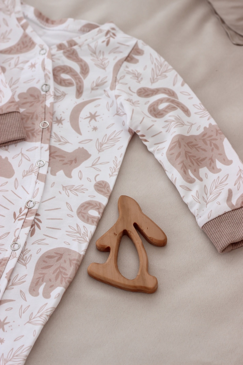May include: A white baby onesie with a brown and white woodland animal print. The onesie has a snap closure and long sleeves. A wooden bunny teether is lying on the onesie.
