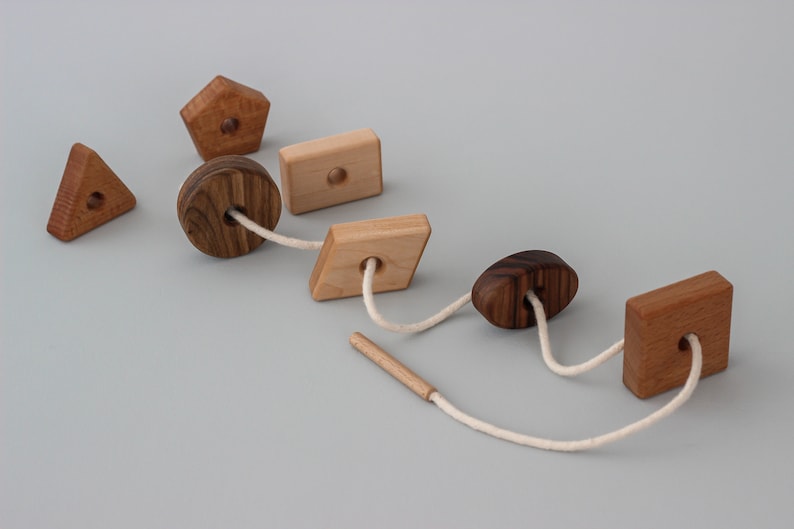 May include: A wooden toy with different shapes strung together with a white cord. The shapes include a triangle, a circle, a rectangle, a diamond, an oval, and a square.