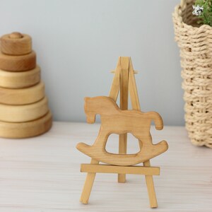 May include: A wooden rocking horse cutout displayed on a small wooden easel. The rocking horse is a light brown colour and the easel is a natural wood colour.