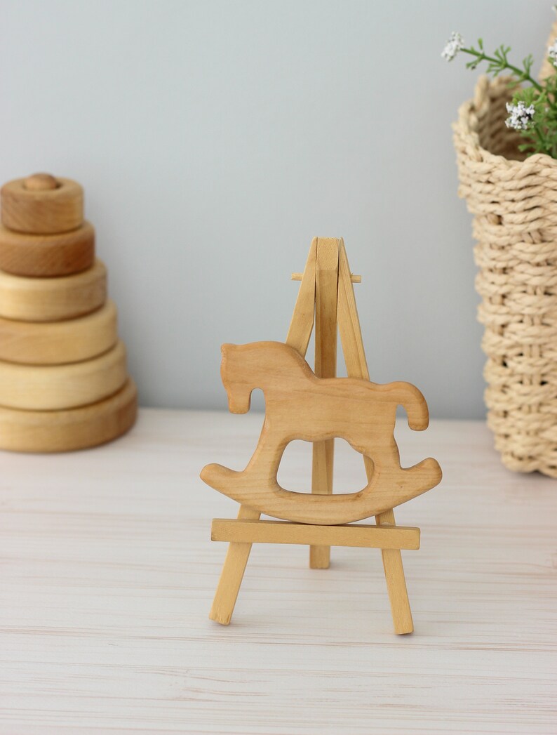 May include: A wooden rocking horse cutout displayed on a small wooden easel. The rocking horse is a light brown colour and the easel is a natural wood colour.