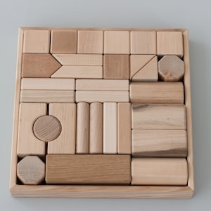Puede incluir: Una caja de madera llena de bloques de madera de diferentes formas y tamaños. Los bloques son todos de color madera natural y están dispuestos en un patrón.