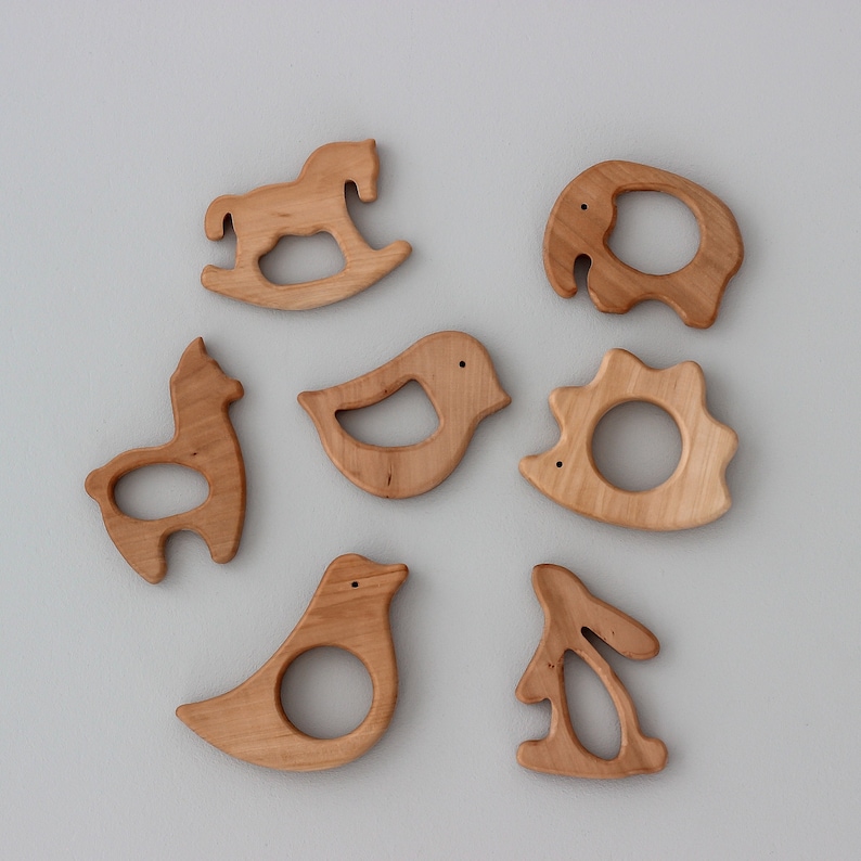 May include: Set of seven wooden teethers shaped like animals, including a rocking horse, bird, llama, hedgehog, elephant, bunny, and another bird. The teethers are made of natural wood and have a smooth, rounded finish.