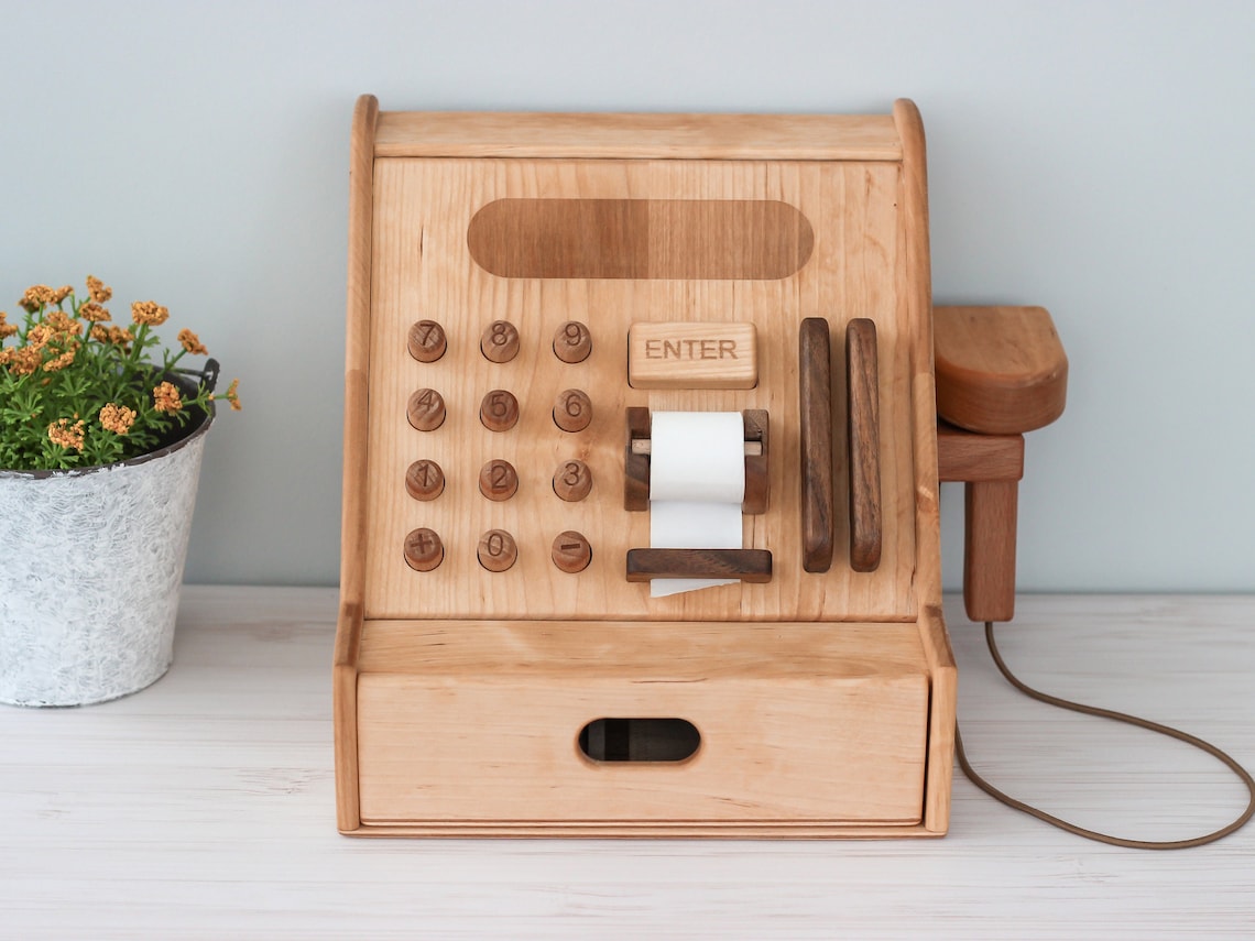 May include: A wooden toy cash register with a drawer, a paper roll, and a number pad with buttons labeled 1 through 9, plus, minus, and enter. The cash register is light brown with a dark brown trim. The cash register has a brown cord attached to it.