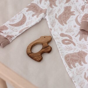 May include: A white baby onesie with a brown bear print and a wooden bear teether. The onesie has a brown trim around the neck and wrists.