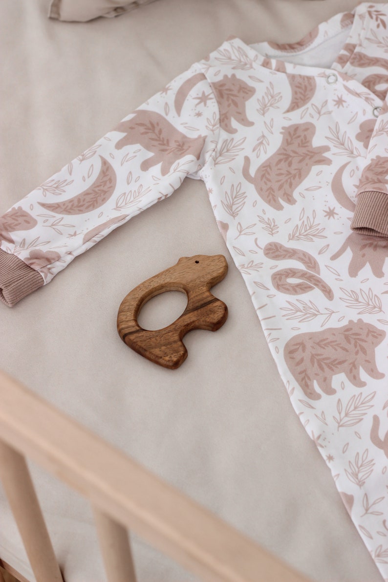 May include: A white baby onesie with a brown bear print and a wooden bear teether. The onesie has a brown trim around the neck and wrists.