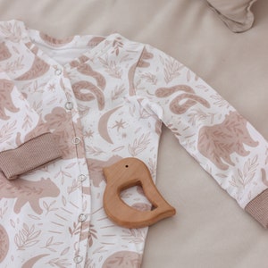May include: A white baby onesie with a brown bear and leaf print. The onesie has a button closure and is lying on a beige surface with a wooden bird-shaped teether.