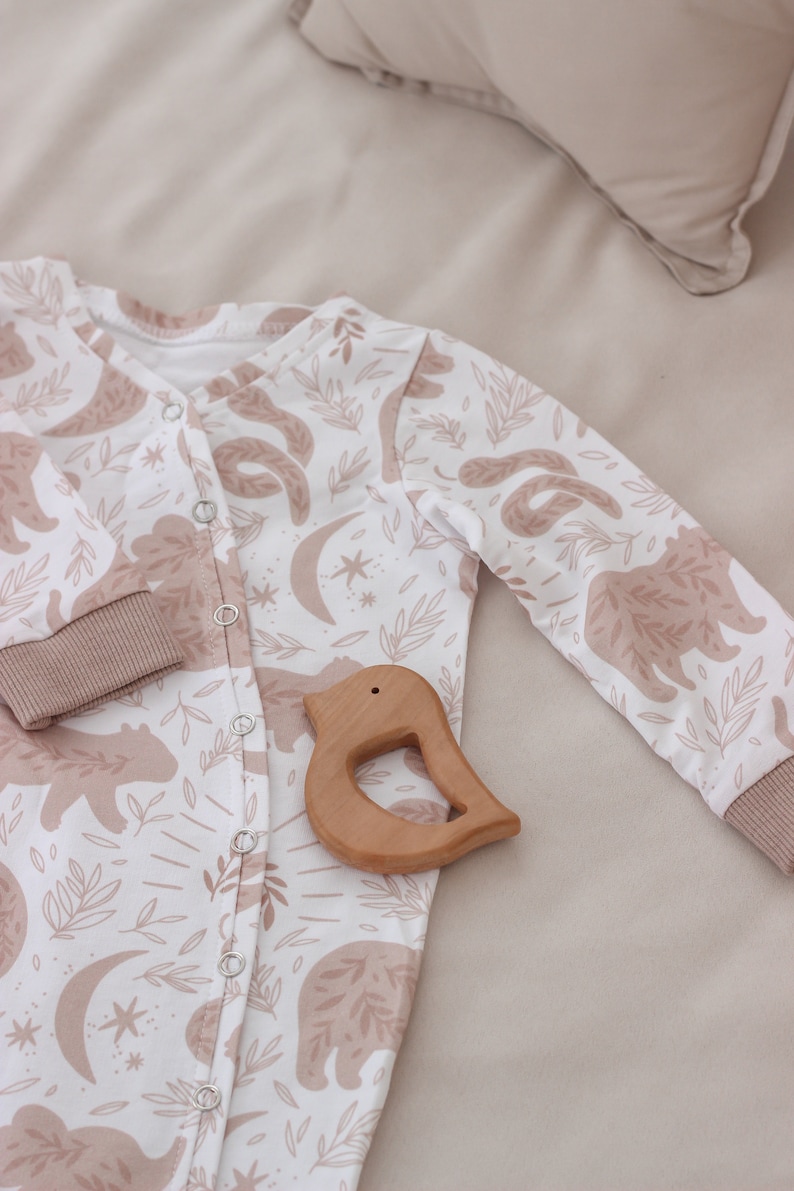 May include: A white baby onesie with a brown bear and leaf print. The onesie has a button closure and is lying on a beige surface with a wooden bird-shaped teether.