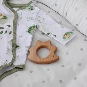 May include: A white baby onesie with a green trim and a leafy print. A wooden teether shaped like a hedgehog is on a white and grey polka dot sheet.