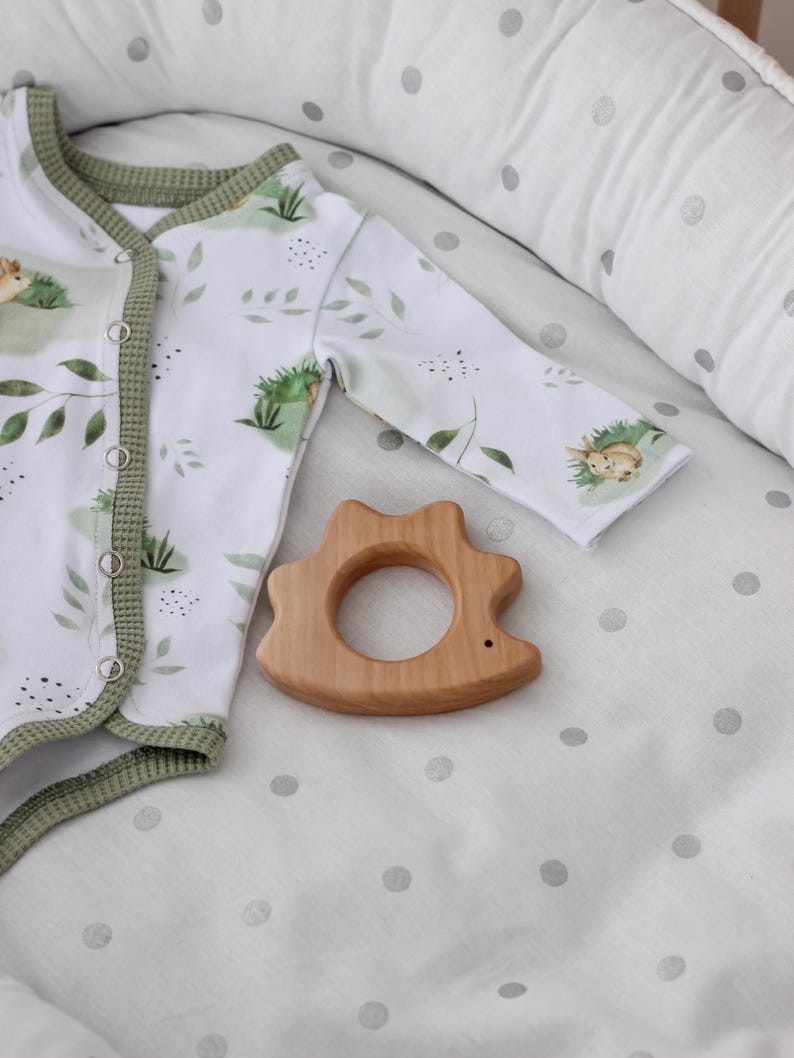 May include: A white baby onesie with a green trim and a leafy print. A wooden teether shaped like a hedgehog is on a white and grey polka dot sheet.