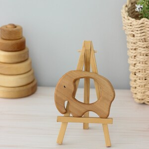 May include: A wooden elephant teether on a small wooden easel. The teether is light brown and has a hole in the middle. The easel is also light brown and has three legs.