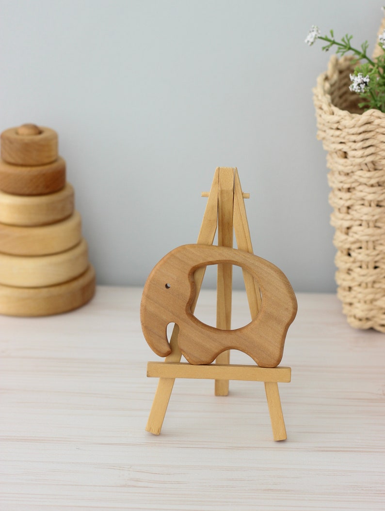 May include: A wooden elephant teether on a small wooden easel. The teether is light brown and has a hole in the middle. The easel is also light brown and has three legs.