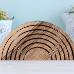 Pode incluir: Um brinquedo de empilhar de madeira em forma de arco-íris com sete arcos de tamanhos diferentes. Os arcos são feitos de madeira natural com uma mancha marrom escura. O brinquedo está disposto em semicírculo sobre uma superfície branca.