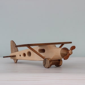 Può includere: Un aereo di legno giocattolo con ali marroni e un'elica. L'aereo ha un corpo marrone chiaro con tre piccoli fori su ogni lato. L'aereo è seduto su una superficie di legno bianco.