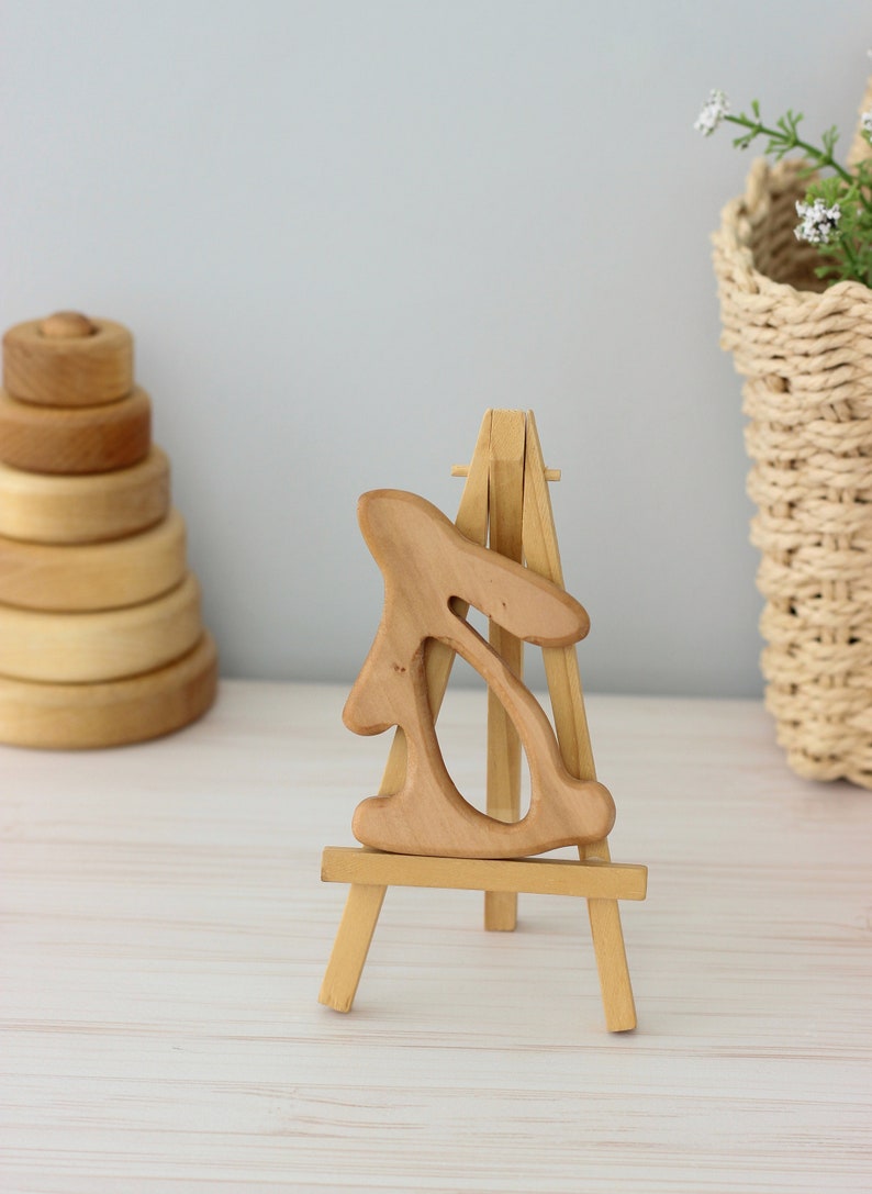 May include: A wooden rabbit sculpture on a small wooden easel. The rabbit is carved in a simple, abstract style and has a light brown colour.
