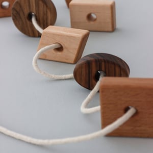 May include: A wooden toy with different shapes strung together with a white cord. The shapes include a circle, a square, a diamond, a pentagon, and an oval. The shapes are made of different types of wood, including walnut and maple.