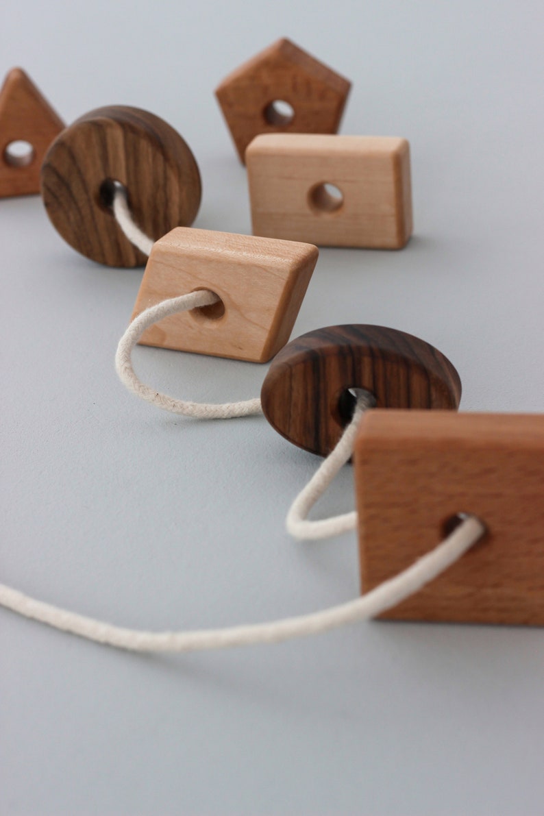 May include: A wooden toy with different shapes strung together with a white cord. The shapes include a circle, a square, a diamond, a pentagon, and an oval. The shapes are made of different types of wood, including walnut and maple.