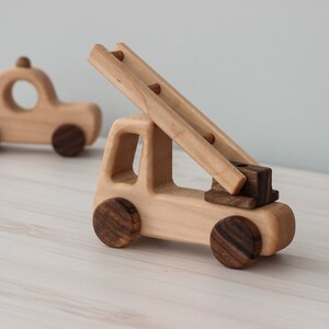 Op de afbeelding: Een houten speelgoed brandweerwagen met een ladder en bruine wielen. De brandweerwagen is gemaakt van licht hout en heeft een eenvoudig ontwerp. Er is een kleinere houten speelgoedauto op de achtergrond.
