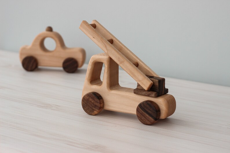 Op de afbeelding: Een houten speelgoed brandweerwagen met een ladder en bruine wielen. De brandweerwagen is gemaakt van licht hout en heeft een eenvoudig ontwerp. Er is een kleinere houten speelgoedauto op de achtergrond.