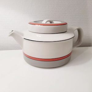 Könnte beinhalten: Eine Keramik-Teekanne mit weißem Körper, grauem Boden und passendem Deckel. Das Design umfasst eine dünne schwarze horizontale Linie und ein breiteres korallenrotes Band, das sowohl den Deckel als auch den unteren Teil des Kannenbauchs umschließt.