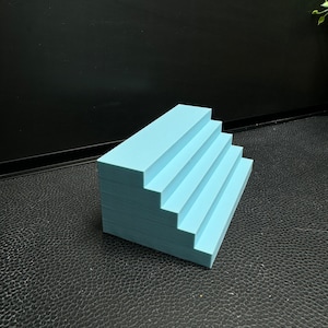 May include: A light blue, five-step staircase made of cardboard. The staircase is sitting on a black textured surface.