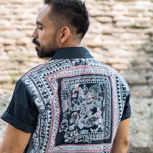 May include: A black short-sleeved shirt with a white and red patterned design featuring an intricate Aztec-inspired motif. The shirt has a button-up collar and a relaxed fit.